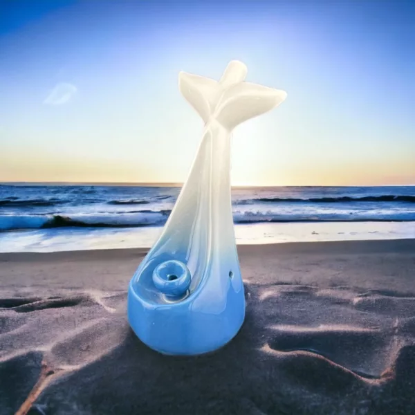 White ceramic dolphin tail pipe with blue tail, captured on a sandy beach at sunset. Perfect for smoking enthusiasts.