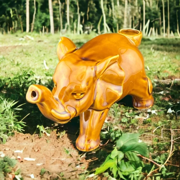 Handcrafted black ceramic elephant pipe with closed eyes and trunk holding something, standing in a green grassy field with trees in the background.