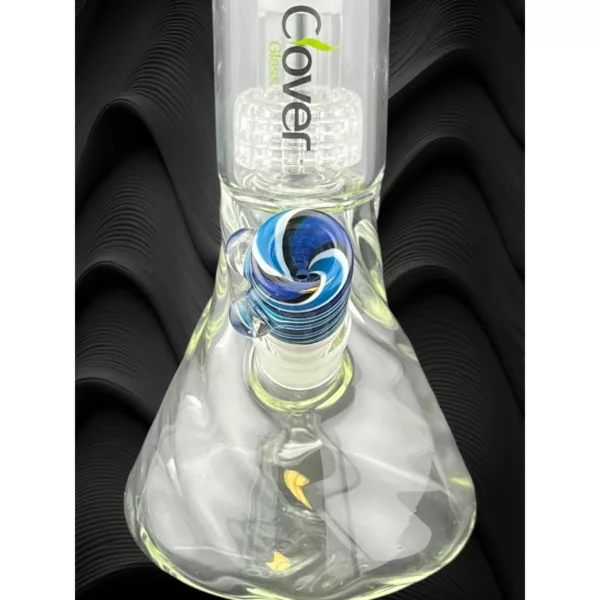 This image showcases the Candy Vortex Clover Bowl, a glass bong with a blue and green swirl design on the base. It features a clear glass stem and base with a small hole at the top and bottom, sitting on a black surface with a wavy pattern. The image is well lit.