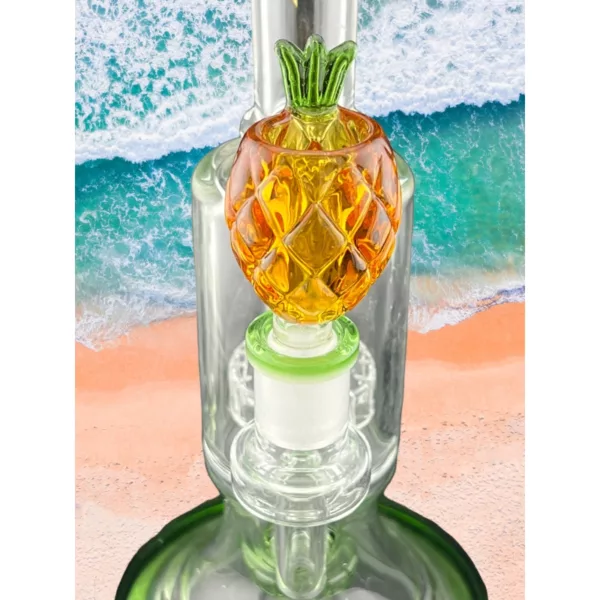 Glass bong with clear body and green stem, featuring a pineapple on top. Set on sandy beach with blue sky and natural light. Wide angle shot with high resolution and natural style.