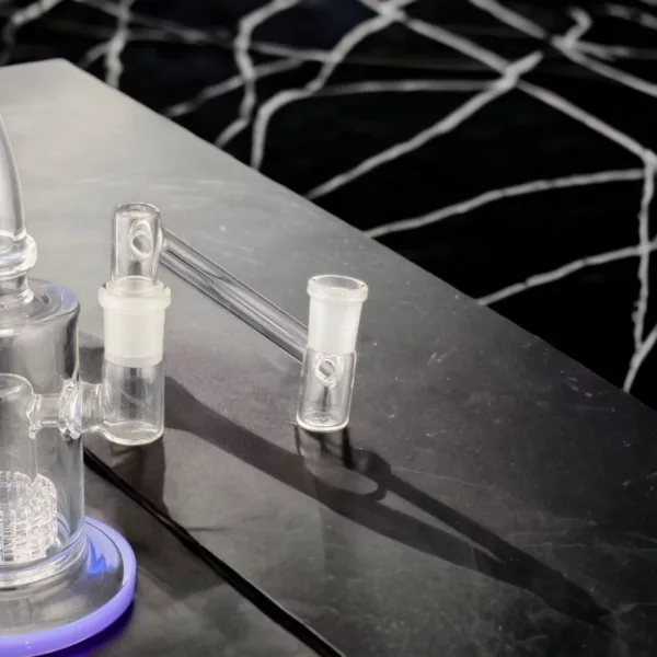 glass bong with a clear, cylindrical shape and two small glass cups. It sits on a black countertop with a checkerboard pattern and is illuminated by a small white light. The atmosphere is dark and mysterious.