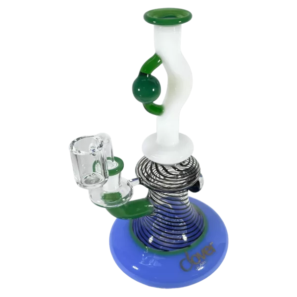 Surreal BDL WP - CCWPE630 features a clear glass waterpipe with a blue and green striped base, a wooden stand with a glass bowl, and a small hole on the side and bottom of the bowl. The bowl has a blue, green, and white pattern and is surrounded by a wooden stand with a blue and green stripe pattern. The large glass tube on the side has a small hole for smoke to be drawn from and a small, clear glass tube attached to the top. The glass tube on the side is filled with water.