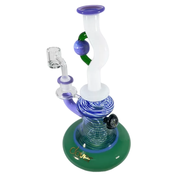 A colorful glass bong with a clear base and bowl, sitting on a stand with a small handhold on the side and a purple/green ring around the middle.