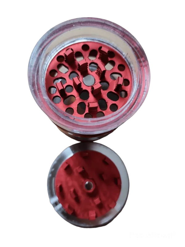 Red metal grinder with clear plastic lid and small hole in center. Circular shape with metal piece on top. Elegant design.