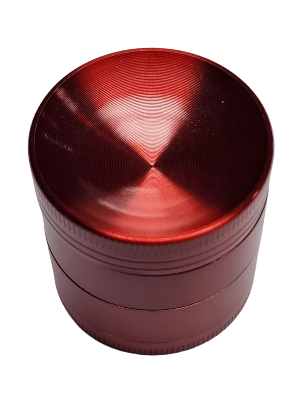 A red, metallic grinder with a flat top and circular base designed for grinding herbs and spices. The base has an embossed pattern and a slot for herbs.